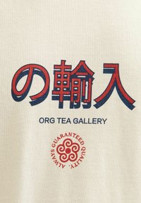 Kreminės spalvos medvilninis marškinėliai su raudona ir mėlyna stilizuota užrašu: „の輸入“ ir „ORG TEA GALLERY“, su raudonu apskrito formos kokybės logotipu.