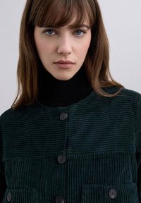 Chaqueta de pana verde oscuro con textura acanalada, que presenta un cuello alto negro tipo mock y botones delanteros, además de dos bolsillos frontales.