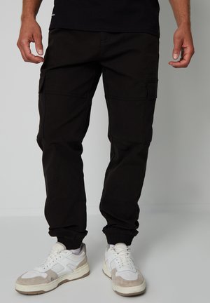 Schwarze Cargo-Hosen aus Baumwolle mit Seitentaschen, elastischen Bündchen und einer lockeren Passform, getragen mit weißen und grauen Sneakers.