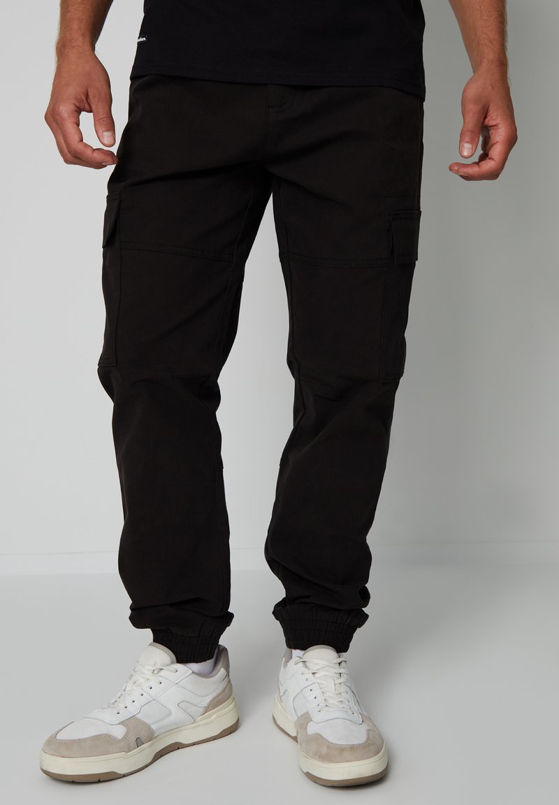 Zwarte cargobroek gemaakt van katoen, met zijzakken, elastische boorden en een relaxte pasvorm, gecombineerd met wit en grijs sneakers.