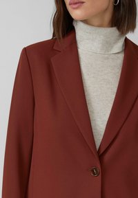 Vrouw in een roestkleurige blazer over een lichtbeige coltrui met subtiele sprankels, met kort bruin haar en een zichtbare oorring.