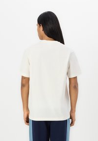 Tricou din bumbac crem, cu mâneci scurte, cu guler rotund, croială lejeră și o textură netedă. Spatele este simplu, fără grafică sau accente.