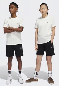 Two children are wearing matching white t-shirts and black shorts. The t-shirts feature a small black logo, and they wear striped socks with sneakers.