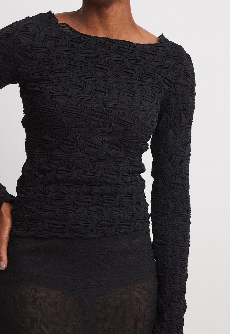 Mujer vistiendo una blusa negra de manga larga texturizada y una falda negra transparente, mostrando detalles de la tela y un estilo ajustado.