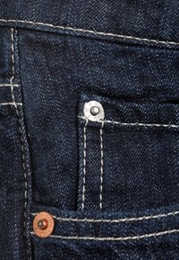 Gros plan d'une poche en jean denim bleu foncé avec des rivets visibles et des coutures blanches sur le tissu.