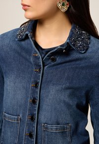 Veste en denim avec une teinture foncée, un col orné, deux poches avant et des boutons noirs le long de la patte de boutonnage. Détails en perles texturées sur le col.