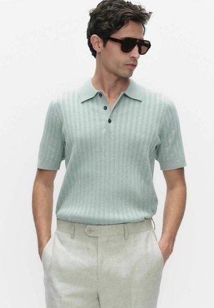 Mężczyzna w jasnozielonej, teksturowanej, krótkorękawowej koszulce polo, jasnobeżowych spodniach i ciemnych okularach przeciwsłonecznych, pozuje z rękami w kieszeniach na białym tle.