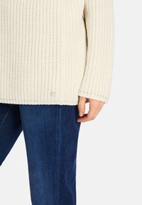 Cream-colored, ribbed knit sweater with wide sleeves, a relaxed fit, and a small metal logo accent near the hem, paired with blue jeans.