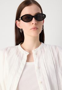 Black cat-eye sunglasses with a thick frame, worn with a sheer white blouse featuring vertical pleats and a round, buttoned collar.