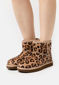 Leopard print ankle boots made of soft suede, featuring a shearling lining, round toe, and a flat rubber sole. Adjustable strap detail.