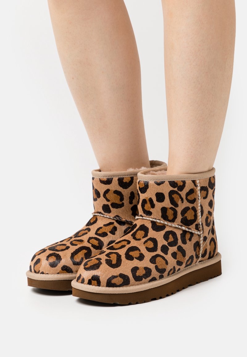 Leopard print ankle boots made of soft suede, featuring a shearling lining, round toe, and a flat rubber sole. Adjustable strap detail.