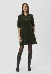 Mujer de pie con un vestido corto de manga corta verde oscuro con cremallera frontal, medias negras y botas de cuero negras hasta la rodilla.