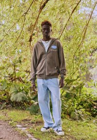 Sweat à capuche marron zippé avec logo, T-shirt blanc en dessous, jean bleu clair, et baskets noires ; debout parmi un feuillage vert.