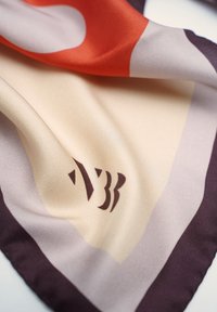 Silk scarf featuring an abstract design in orange, beige, and purple, with a smooth texture and a dark logo on one edge.