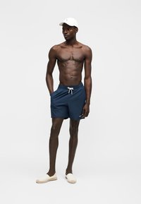 Άνδρας χωρίς πουκάμισο που φοράει λευκό καπέλο, σκούρα μπλε σορτς με κορδόνι και λευκά παπούτσια χωρίς κορδόνια, στέκεται με τα χέρια στις τσέπες.