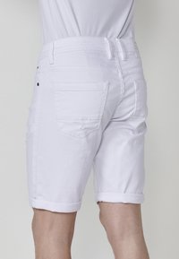 Pantalones cortos de denim blanco con dobladillo enrollado, que presentan dos bolsillos traseros y un sutil desgastado, sobre un fondo gris claro.