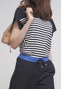 Woman wearing navy and white striped shirt and navy pants with blue waistband, holding tan perforated leather backpack over shoulder.