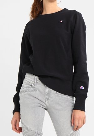 Femme portant un sweat-shirt noir Champion avec logos sur la poitrine et la manche, associé à un jean gris clair sur fond uni.