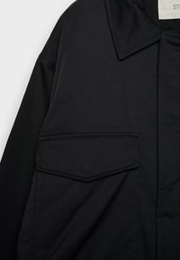 Sort jakke med spids krave, stor frontlomme og glat stof med synlige sømdetaljer.