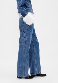 Wijde jeans van middelblauwe denim met grote zakken, vervaagde textuur en beige stiksels. Gecombineerd met een witte faux fur accent.