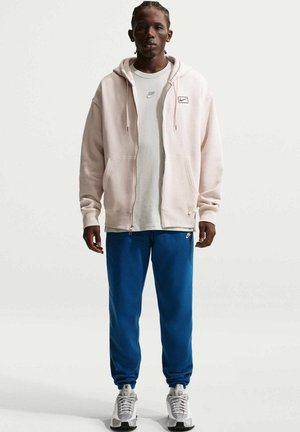 Ljusrosa zip-up-hoodie med framficka, vit t-shirt och blåa mjukisbyxor. Sneakers med vita och svarta detaljer. Enkel design.