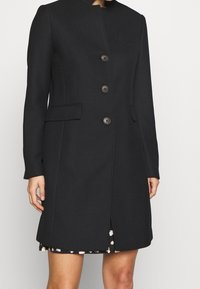 Black coat made of textured fabric, featuring a high collar, front button closure, and two side pockets. Mid-length with a slit at the back.