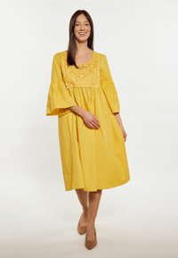 Gelbes Baumwolkleid mit quadratischem Ausschnitt und bestickten Details, mit Glockenärmeln und fließender Silhouette.
