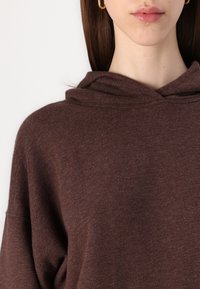 Smeđa dukserica s kapuljačom, mekane teksture, opuštenog kroja, širokog izreza i spuštenih ramena. Minimalistički dizajn, bez vidljivih uzoraka ili akcenta.