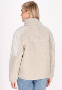 Giacca in fleece color crema con un corpo morbido e texturizzato e pannelli spalla lisci di colore chiaro; presenta un colletto alto e una vestibilità ampia.