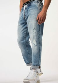 Pantalones jogger de denim azul claro con detalles desgastados, cintura elástica con cordón y puños, combinados con zapatillas deportivas blancas.