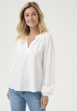 White long-sleeve blouse with a V-neckline and slightly gathered sleeves. Made of lightweight textured fabric, paired with blue jeans.