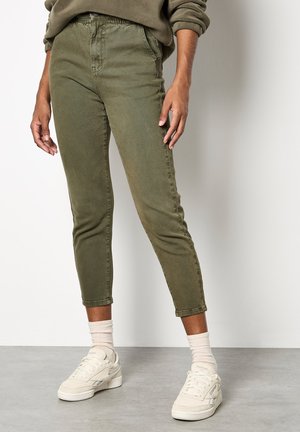 Pantaloni cropped in misto cotone verde oliva con design fitted, dotati di tasche frontali e texture morbida, abbinati a sneakers bianche e calze leggere.