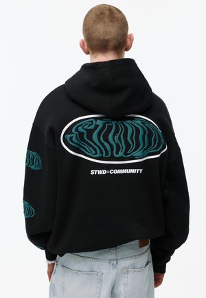 Osoba s kratkom kosom u crnoj dukserici s kapuljačom na kojoj se na leđima i rukavima nalazi tirkizni iskrivljeni tekst "STWD" i "STWD COMMUNITY".