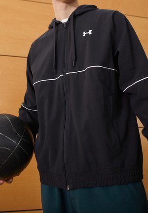 Osoba v černé mikině na zip značky Under Armour držící černý basketbalový míč na pozadí dřevěné stěny.