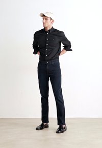 Black button-up shirt with collar, dark jeans, and black shoes. Tan cap on head. Model stands with hands behind back against white background.