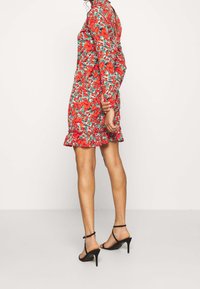 Rotes Blumen Kleid mit langen Ärmeln, Rüschensaum und lebhaftem grünem Laub. Getragen mit schwarzen Riemchen-Heels. Das Muster zeigt große Blüten.