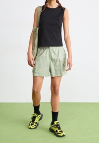 Top negro sin mangas, pantalones cortos verde claro y calcetines negros. Lleva zapatillas en amarillo y gris, con un bolso de mano verde colgado al hombro.