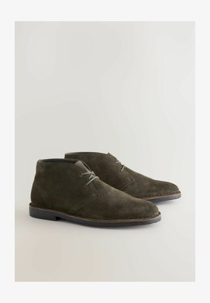 Temno olivno zelene ankel čizme iz sredine zatezanja z meko teksturo, zaobljenim prstom in minimalnim vezanjem. Lahka gumijasta podplata in preprost dizajn.