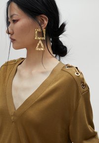 Top a maglia color senape con scollatura a V e dettagli ad occhiello decorativi sulle spalle; grandi orecchini geometrici in oro con texture martellata.