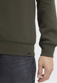 Felpa verde oliva con orlo e polsini a costine, con un piccolo etichetta nera in fondo. Jeans con una finitura scura e testurizzata visibile.