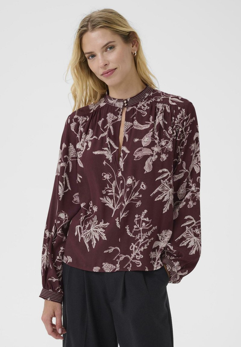Kvindelig med langt, bølget hår iført en maroon bluse med blomstermønster og en nøglehulåbning, parret med sorte bukser, smiler blidt.