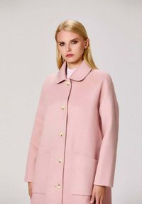Manteau en laine rose avec un col arrondi, fermeture à boutons sur le devant, deux grandes poches et une finition douce et texturée.