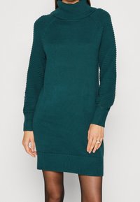 Robes pull en tricot teal avec un col haut, de longues manches en tricot présentant des accents texturés, et des poignets et un ourlet côtelés. Fini en tissu lisse.