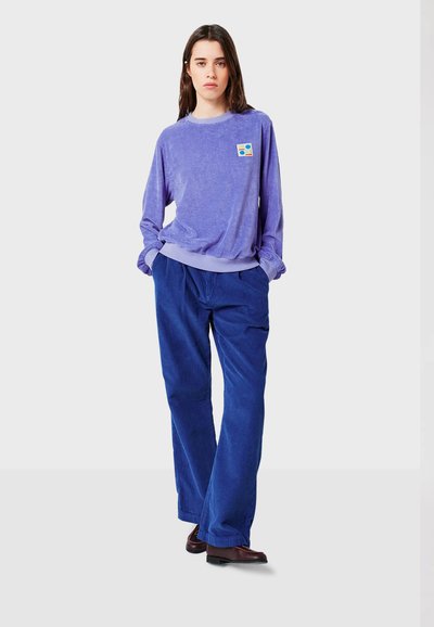 Camicetta viola a maniche lunghe con maniche risvoltate e piccolo logo, abbinata a pantaloni in corduroy blu a gamba larga. Texture liscia e vestibilità rilassata.