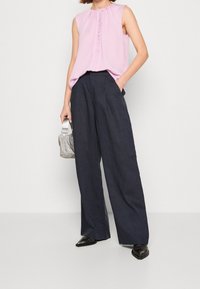 Top sin mangas rosa con volantes en el frente, combinado con pantalones de pierna ancha en azul marino oscuro. Un bolso texturizado plateado completa el conjunto. Zapatos negros de punta.