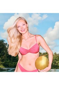 Rosa bikini med rynkdetaljer, kombinerad med en matchande nederdel. Modellen håller en gyllene kokosnöt, mot en blå himmel och frodig grönska.
