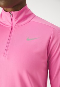 Růžový top Nike s půlvýstřihem na zip, vyrobený z hladké, prodyšné látky s vysokým límcem. Na levé straně hrudi je malý stříbrný logo Nike.
