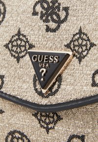Borsa a tracolla in tessuto beige con motivo floreale nero, dotata di una placca triangolare nera e oro con la scritta "GUESS USA".