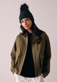 Veste vert olive avec un col marron et deux poches avant, portée sur une chemise noire, complétée par un bonnet en tricot noir avec un pompon.
