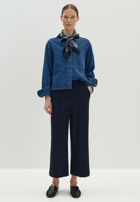 Blauwe spijkerjack met kraag en knoopsluiting, gecombineerd met ruime marineblauwe broek. Zwarte sjaal om de nek geknoopt en zwarte instappers.